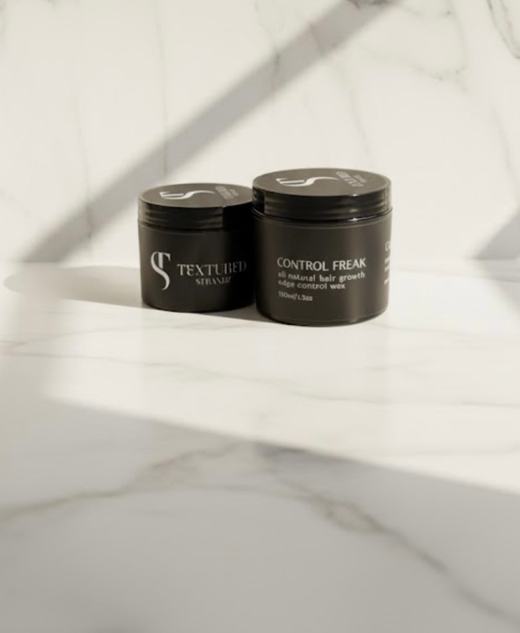 Two black hair product jars labeled 'Textured' and 'Control Freak' on a marble surface.